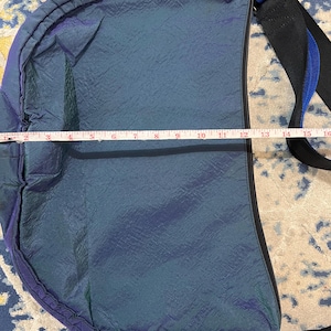 K&ouml;nnte beinhalten: Blau-violette, schillernde Umh&auml;ngetasche mit schwarzem Rei&szlig;verschluss und Riemen. Die Tasche ist ca. 38 cm lang. Der Riemen ist schwarz mit blauem Streifen. Die Tasche hat eine strukturierte Oberfl&auml;che.