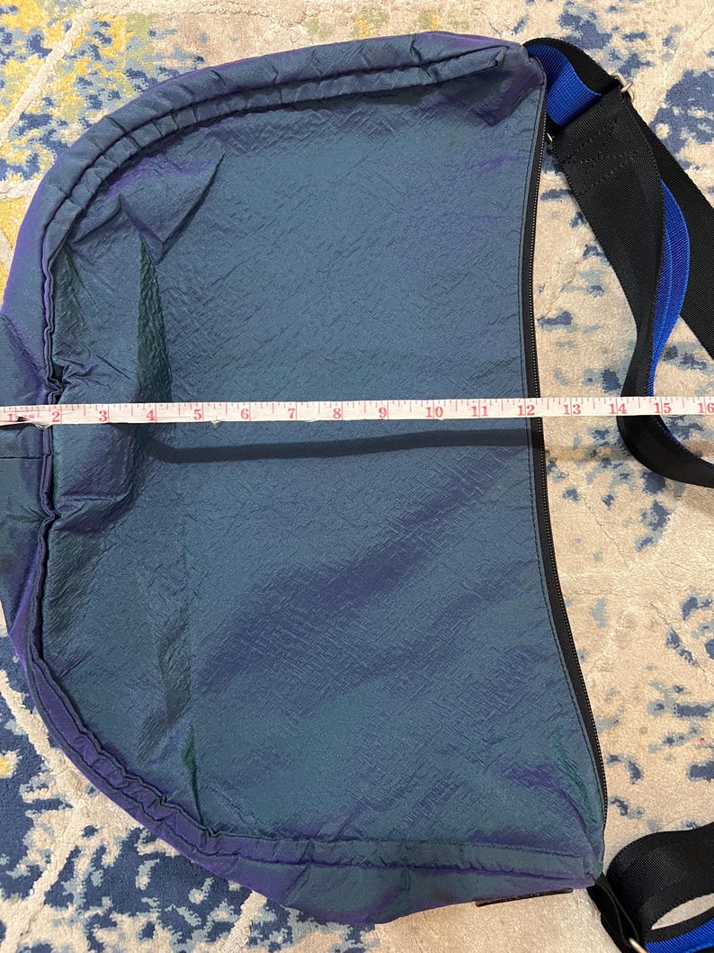 K&ouml;nnte beinhalten: Blau-violette, schillernde Umh&auml;ngetasche mit schwarzem Rei&szlig;verschluss und Riemen. Die Tasche ist ca. 38 cm lang. Der Riemen ist schwarz mit blauem Streifen. Die Tasche hat eine strukturierte Oberfl&auml;che.