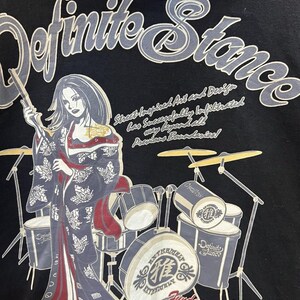 Pu&ograve; includere: T-shirt nera con grafica di una donna in kimono con bacchette, con una batteria sullo sfondo. Visibili anche le scritte "Definite Stance" e "Fundamental Recurrence".