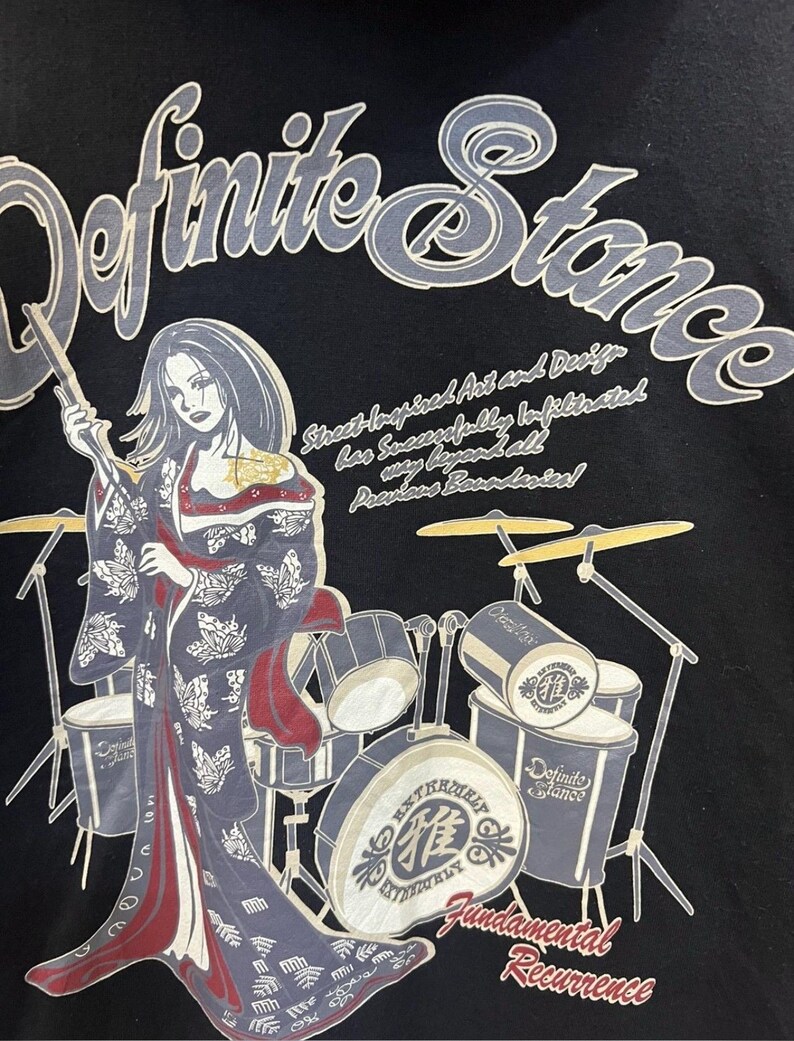 Pu&ograve; includere: T-shirt nera con grafica di una donna in kimono con bacchette, con una batteria sullo sfondo. Visibili anche le scritte "Definite Stance" e "Fundamental Recurrence".