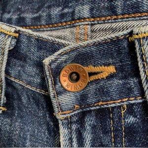 Peut inclure: Gros plan sur un jean en denim bleu avec un bouton cuivr&eacute;. Le bouton porte l'inscription "EDWIN". Le jean pr&eacute;sente des coutures jaunes et une fermeture boutonn&eacute;e.