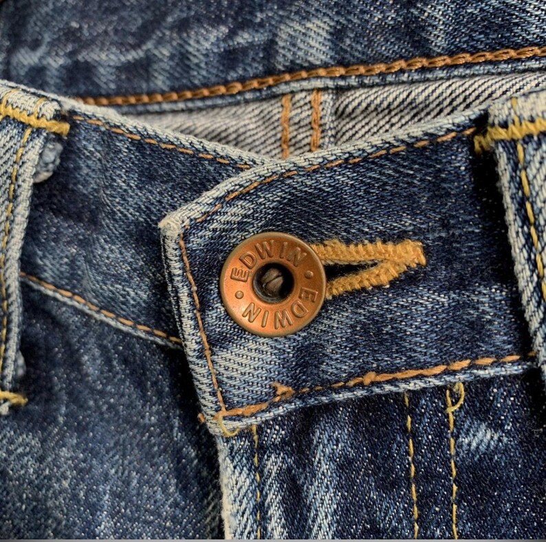 Peut inclure: Gros plan sur un jean en denim bleu avec un bouton cuivr&eacute;. Le bouton porte l'inscription "EDWIN". Le jean pr&eacute;sente des coutures jaunes et une fermeture boutonn&eacute;e.