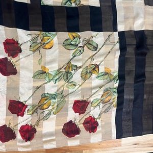 May include: Silk scarf with a floral rose pattern. The scarf features red roses, green leaves, and beige and black stripes. The scarf has a label in the corner.
