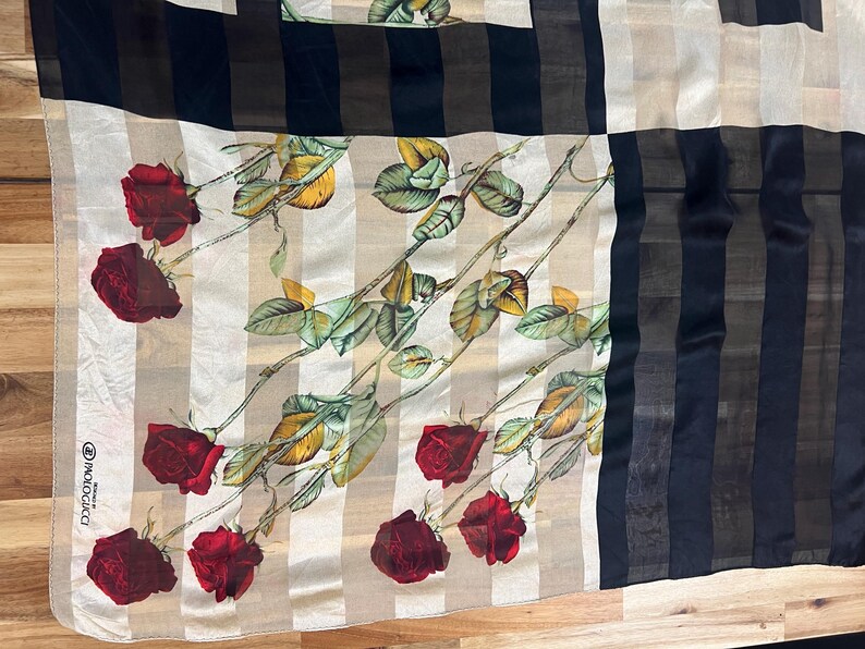 May include: Silk scarf with a floral rose pattern. The scarf features red roses, green leaves, and beige and black stripes. The scarf has a label in the corner.