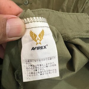 May include: An olive green garment featuring a white fabric tag with the Avirex logo, which displays a gold winged star emblem above the brand name. The tag includes Japanese text regarding garment care instructions and color bleeding warnings.