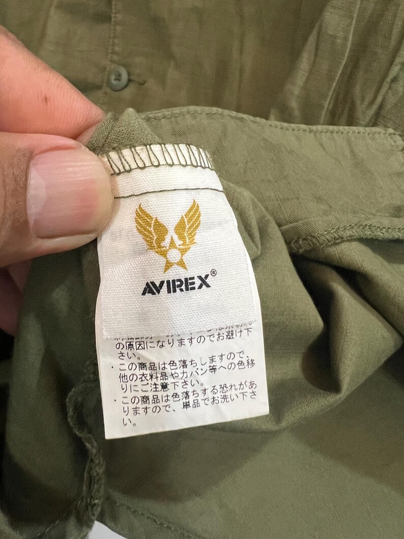 May include: An olive green garment featuring a white fabric tag with the Avirex logo, which displays a gold winged star emblem above the brand name. The tag includes Japanese text regarding garment care instructions and color bleeding warnings.