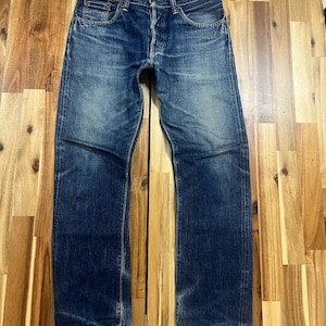 Op de afbeelding: Een paar versleten blauwe spijkerbroeken met een knoopsluiting en een klassiek ontwerp met vijf zakken. De jeans vertonen vervaging en slijtage, met een rechte pijp. De denim heeft een medium wassing met lichtere plekken op de dijen en knieën.