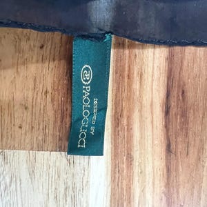 May include: A dark green tag with the text "Paolo Gucci Designed By" in white lettering. The tag is attached to a dark blue fabric, set against a wooden surface.