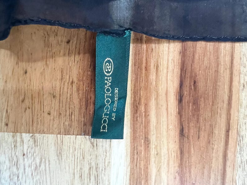 May include: A dark green tag with the text "Paolo Gucci Designed By" in white lettering. The tag is attached to a dark blue fabric, set against a wooden surface.