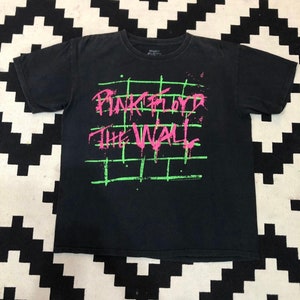 Peut inclure: T-shirt noir avec un imprimé graphique rose et vert de "Pink Floyd The Wall".