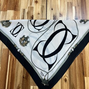 May include: White and black silk scarf with a repeating pattern of interlocking black letter Cs, rope, and decorative elements. The scarf has a black border and the words "CARTIER PARIS".