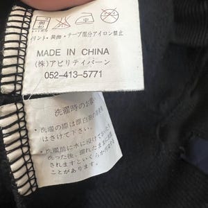 Pu&ograve; includere: Primo piano di un'etichetta di abbigliamento nera con testo giapponese e simboli di cura. L'etichetta recita "MADE IN CHINA" e include un numero di telefono. Sono stampate anche le istruzioni di lavaggio.