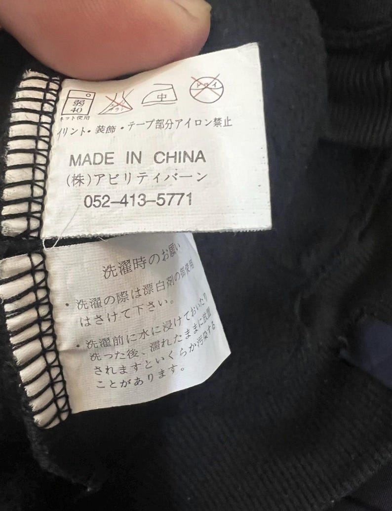 Pu&ograve; includere: Primo piano di un'etichetta di abbigliamento nera con testo giapponese e simboli di cura. L'etichetta recita "MADE IN CHINA" e include un numero di telefono. Sono stampate anche le istruzioni di lavaggio.