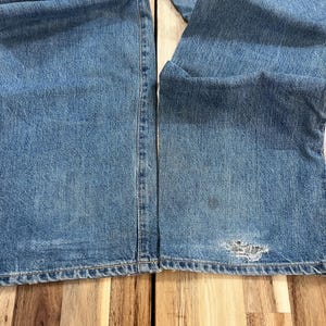 May include: Light blue denim jeans with a distressed area near the knee. The jeans are folded, showing the texture and color variations of the denim. The stitching is visible along the seams.