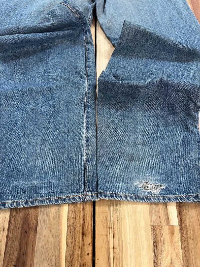 May include: Light blue denim jeans with a distressed area near the knee. The jeans are folded, showing the texture and color variations of the denim. The stitching is visible along the seams.