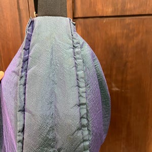 K&ouml;nnte beinhalten: Nahaufnahme einer Umh&auml;ngetasche mit schimmerndem, irisierendem Stoff in Blau-, Gr&uuml;n- und Lilat&ouml;nen. Die Tasche hat einen schwarzen Riemen und eine rechteckige Schnalle. Das Material der Tasche hat ein strukturiertes Aussehen.