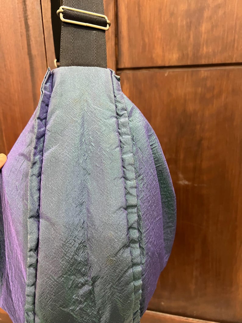 K&ouml;nnte beinhalten: Nahaufnahme einer Umh&auml;ngetasche mit schimmerndem, irisierendem Stoff in Blau-, Gr&uuml;n- und Lilat&ouml;nen. Die Tasche hat einen schwarzen Riemen und eine rechteckige Schnalle. Das Material der Tasche hat ein strukturiertes Aussehen.