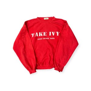 May include: A red sweatshirt with the text "TAKE IVY" in white block letters. Below the text is "GOOD VINTAGE TASTE" in smaller white letters. The sweatshirt is a solid red color.