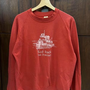 Op de afbeelding: Een rode sweatshirt met een witte afbeelding van een boot en de tekst "Red Stack San Francisco". De crew neck sweatshirt hangt aan een houten hanger.