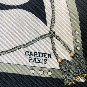 May include: A silk scarf with a striped pattern in black, white, and olive green. The scarf features a rope design with tassels and the text "CARTIER PARIS". The scarf has a pleated texture.