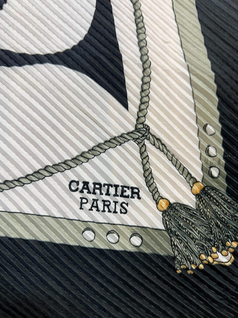 May include: A silk scarf with a striped pattern in black, white, and olive green. The scarf features a rope design with tassels and the text "CARTIER PARIS". The scarf has a pleated texture.