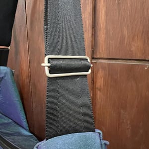 K&ouml;nnte beinhalten: Nahaufnahme eines schwarzen Nylon-Schultergurts mit einer silberfarbenen Metallschnalle. Der Gurt ist an einer t&uuml;rkis-violett schillernden Tasche befestigt. Der Hintergrund ist eine Holzt&uuml;r.