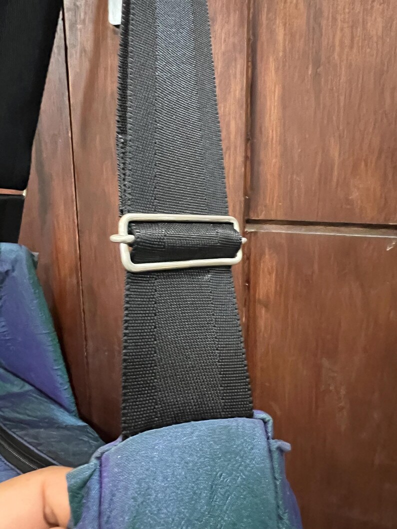 K&ouml;nnte beinhalten: Nahaufnahme eines schwarzen Nylon-Schultergurts mit einer silberfarbenen Metallschnalle. Der Gurt ist an einer t&uuml;rkis-violett schillernden Tasche befestigt. Der Hintergrund ist eine Holzt&uuml;r.