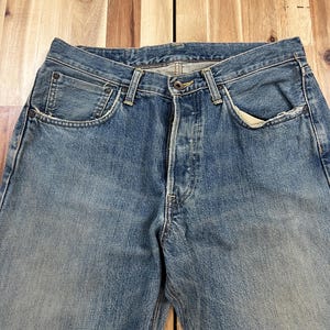 May include: Faded blue denim jeans with classic five-pocket styling. The jeans have a button fly and show signs of wear, with lighter areas on the thighs and knees. The fabric appears to be a medium weight.