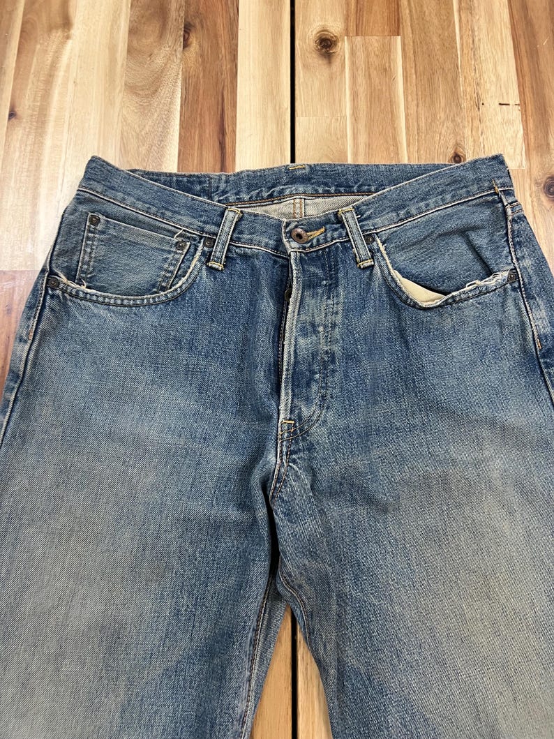 May include: Faded blue denim jeans with classic five-pocket styling. The jeans have a button fly and show signs of wear, with lighter areas on the thighs and knees. The fabric appears to be a medium weight.
