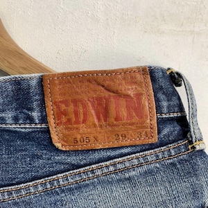 Peut inclure: Gros plan sur un jean en denim bleu avec un patch en cuir marron. Le patch pr&eacute;sente le nom de la marque "EDWIN" en lettres rouges et le texte "505 X 29 34". Le jean a des coutures visibles.