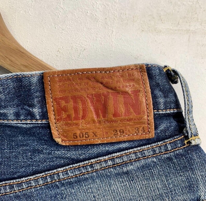 Peut inclure: Gros plan sur un jean en denim bleu avec un patch en cuir marron. Le patch pr&eacute;sente le nom de la marque "EDWIN" en lettres rouges et le texte "505 X 29 34". Le jean a des coutures visibles.