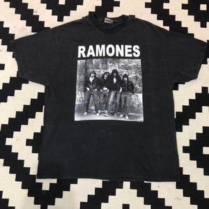 Peut inclure: T-shirt noir avec le nom du groupe "RAMONES" en blanc. En dessous, une photo en noir et blanc des membres du groupe. Le t-shirt est en tissu sombre.