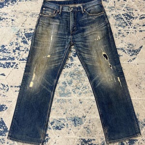 Op de afbeelding: Een blauwe spijkerbroek van denim met een versleten afwerking, vervaagde kreukpatronen, lichtbruine vlekken en verschillende kleine gerafelde scheuren en gaten langs de voorpijpen, compleet met een knoopsluiting en vijf zakken.