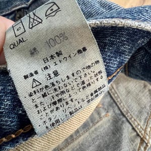 May include: Close-up of a denim garment tag with Japanese text and care symbols. The tag reads "100%" and "Made in Japan." The tag is attached to blue denim fabric.