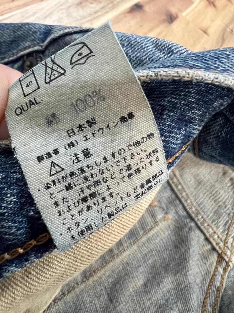 May include: Close-up of a denim garment tag with Japanese text and care symbols. The tag reads "100%" and "Made in Japan." The tag is attached to blue denim fabric.