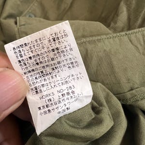 May include: An olive green garment featuring a white rectangular care label sewn into the interior seam. The label contains Japanese text detailing washing instructions, the product code WORKS NO-283, and manufacturer information.