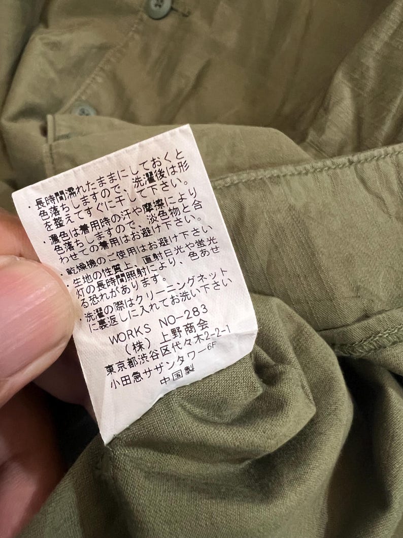 May include: An olive green garment featuring a white rectangular care label sewn into the interior seam. The label contains Japanese text detailing washing instructions, the product code WORKS NO-283, and manufacturer information.