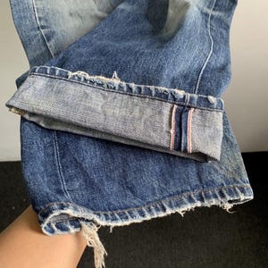 Peut inclure: Gros plan sur un jean en denim bleu avec un aspect us&eacute; et vieilli. Le revers est retrouss&eacute;, r&eacute;v&eacute;lant un bord brut avec une bande rose. Le jean a une conception classique &agrave; cinq poches et un d&eacute;lavage.