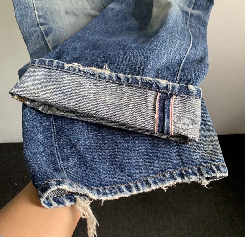 Peut inclure: Gros plan sur un jean en denim bleu avec un aspect us&eacute; et vieilli. Le revers est retrouss&eacute;, r&eacute;v&eacute;lant un bord brut avec une bande rose. Le jean a une conception classique &agrave; cinq poches et un d&eacute;lavage.