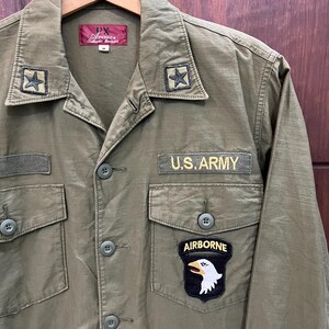 May include: An olive green long-sleeve shirt featuring a U.S. ARMY patch, an AIRBORNE patch with an eagle emblem, and two star patches on the collar. The shirt has a button-down front, two chest pockets with button flaps, and a PX Avirex label.
