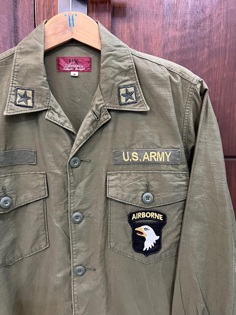 May include: An olive green long-sleeve shirt featuring a U.S. ARMY patch, an AIRBORNE patch with an eagle emblem, and two star patches on the collar. The shirt has a button-down front, two chest pockets with button flaps, and a PX Avirex label.