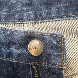 Może przedstawiać: Zbliżenie na ciemnoniebieskie dżinsy. Widoczny jest mosiężny guzik z nazwą marki "Wrangler", a także zamek błyskawiczny i kontrastowe szwy. Tkanina ma teksturowany wygląd.