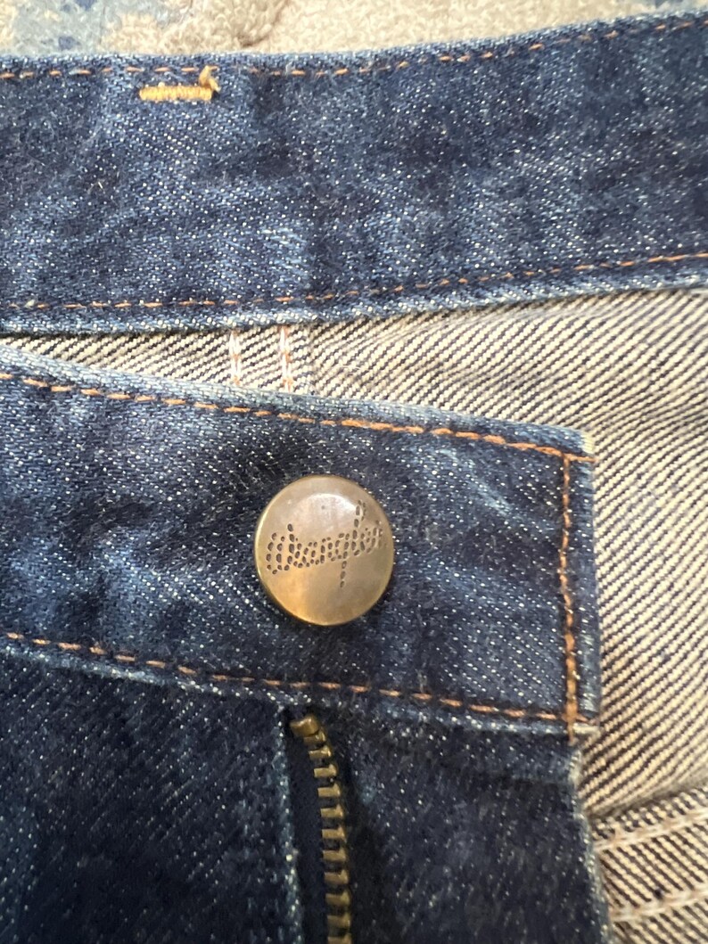 Może przedstawiać: Zbliżenie na ciemnoniebieskie dżinsy. Widoczny jest mosiężny guzik z nazwą marki "Wrangler", a także zamek błyskawiczny i kontrastowe szwy. Tkanina ma teksturowany wygląd.