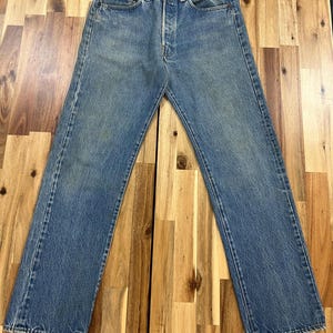 May include: Faded blue denim jeans with a button fly and classic five-pocket design. The jeans show signs of wear and fading, with a straight leg cut. The jeans are laid flat on a wooden surface.