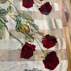 May include: A sheer scarf with a striped pattern and a floral design featuring red roses and green leaves. The scarf is labeled "DESIGNED BY PAOLOGUCCI".