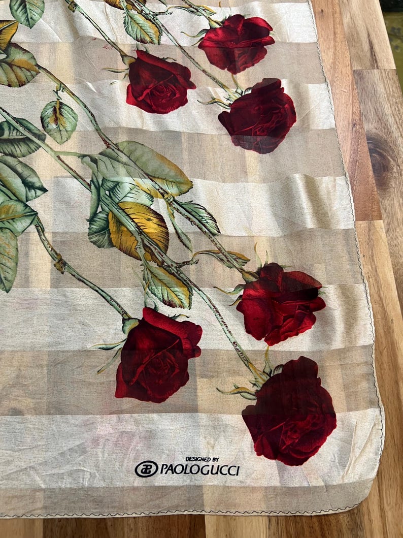 May include: A sheer scarf with a striped pattern and a floral design featuring red roses and green leaves. The scarf is labeled "DESIGNED BY PAOLOGUCCI".
