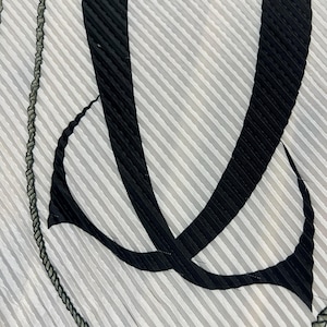 May include: A close-up of a pleated fabric with a striped pattern in shades of gray and white. Bold black abstract shapes and a dark rope-like trim border the design. The fabric has a textured appearance.
