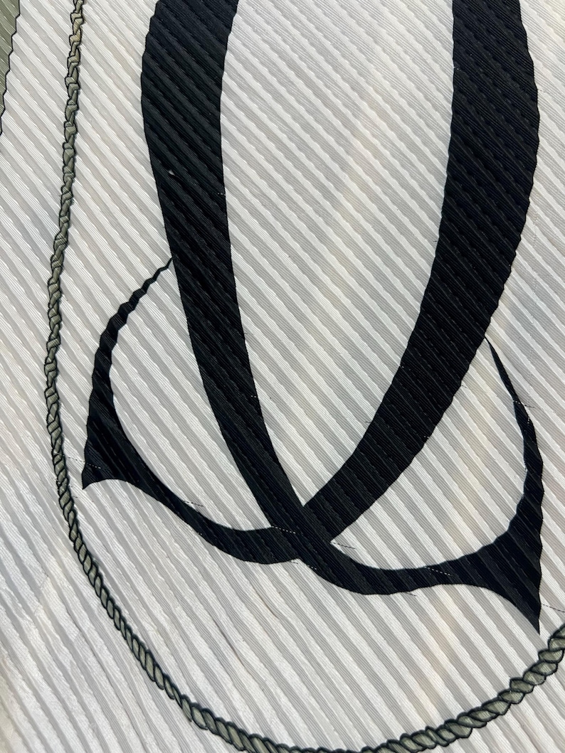May include: A close-up of a pleated fabric with a striped pattern in shades of gray and white. Bold black abstract shapes and a dark rope-like trim border the design. The fabric has a textured appearance.