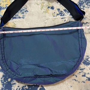 K&ouml;nnte beinhalten: Eine teal- und lilafarbene Umh&auml;ngetasche mit einem schwarzen Riemen und blauen Akzenten. Die Tasche ist etwa 51 cm lang. Die Tasche hat eine gebogene Form und einen Rei&szlig;verschluss. Der Riemen ist verstellbar.