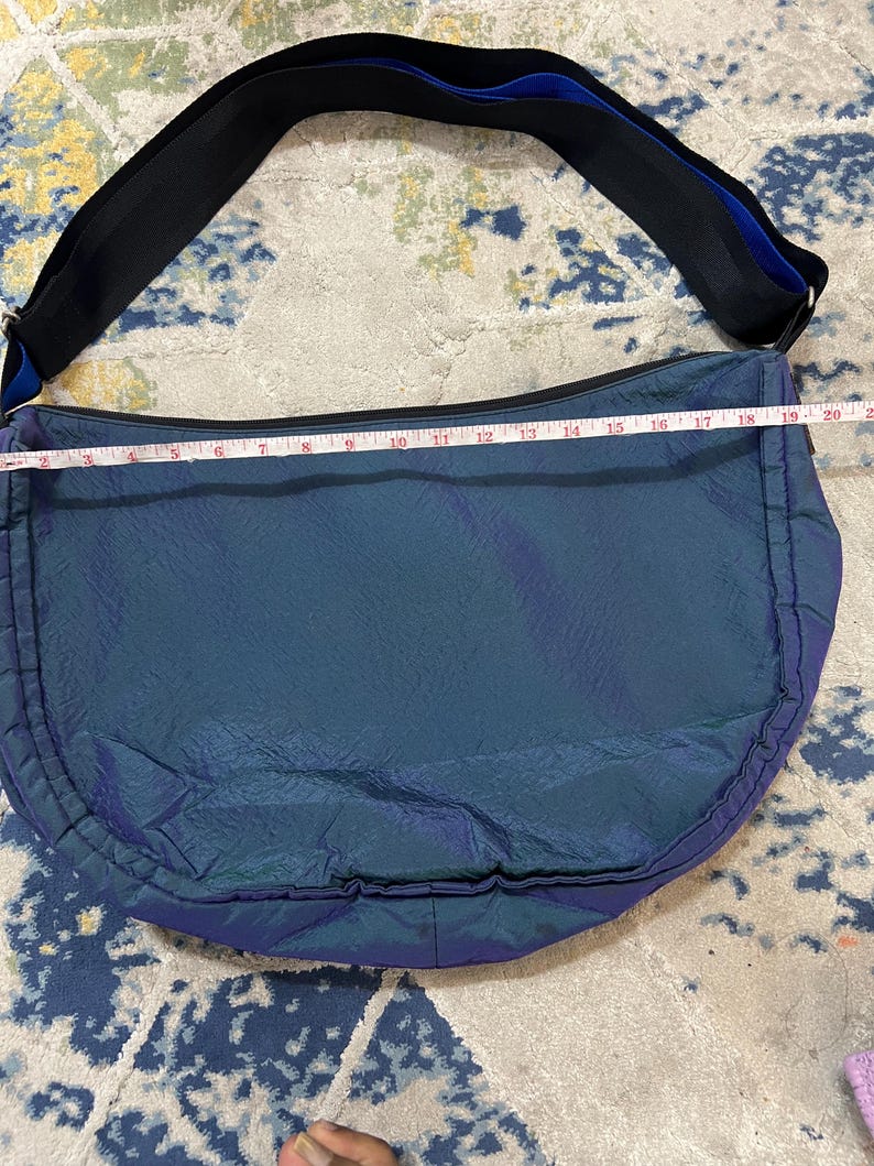 K&ouml;nnte beinhalten: Eine teal- und lilafarbene Umh&auml;ngetasche mit einem schwarzen Riemen und blauen Akzenten. Die Tasche ist etwa 51 cm lang. Die Tasche hat eine gebogene Form und einen Rei&szlig;verschluss. Der Riemen ist verstellbar.